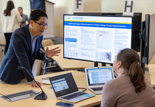 Student stands next to TV monitor showing scholarly project and gestures to woman sitting at table