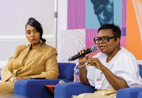 Woman sitting in blue chair holds microphone while speaking to crowd while another woman looks on