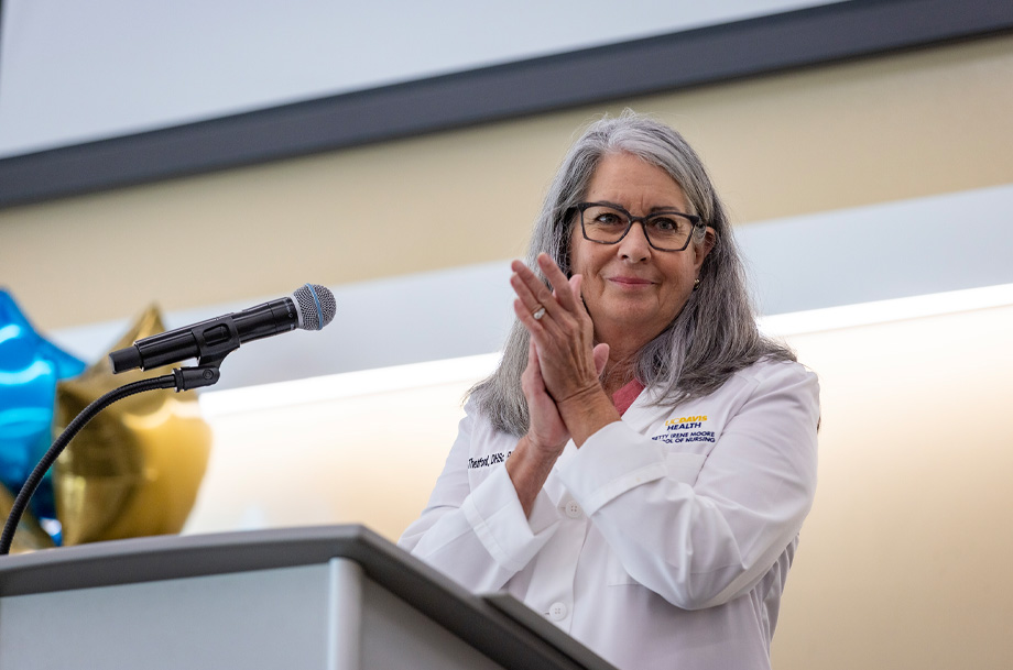 A woman in a white coat stands at a podium with a microphone, clapping and smiling toward the audience during a ceremony.