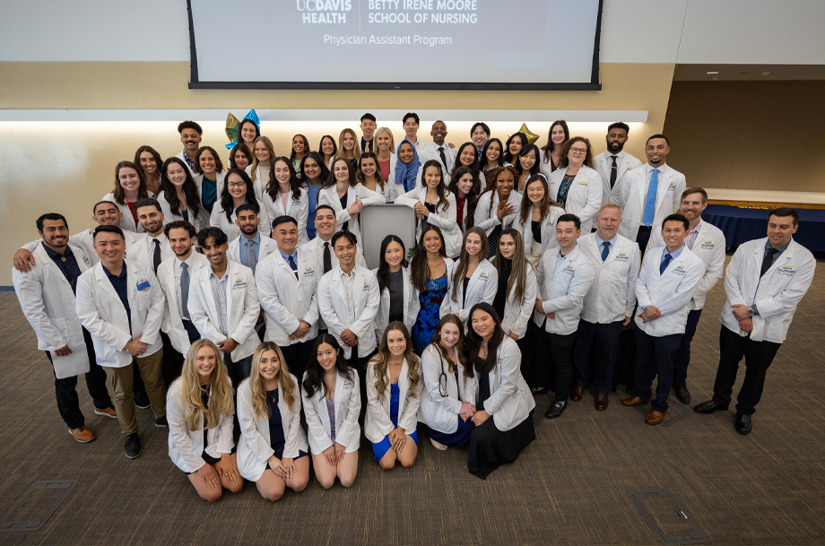 A large group of students in white coats pose together for a class photo at a ceremony.