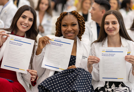 Three smiling students in white coats hold up papers titled ‘Physician Assistant Professional Oath’ during a ceremony, with other classmates seated be