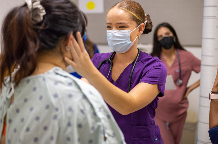 A medical student in purple scrubs and surgical mask uses her left hand to examine a fellow student&rsquo;s face in clinical practice