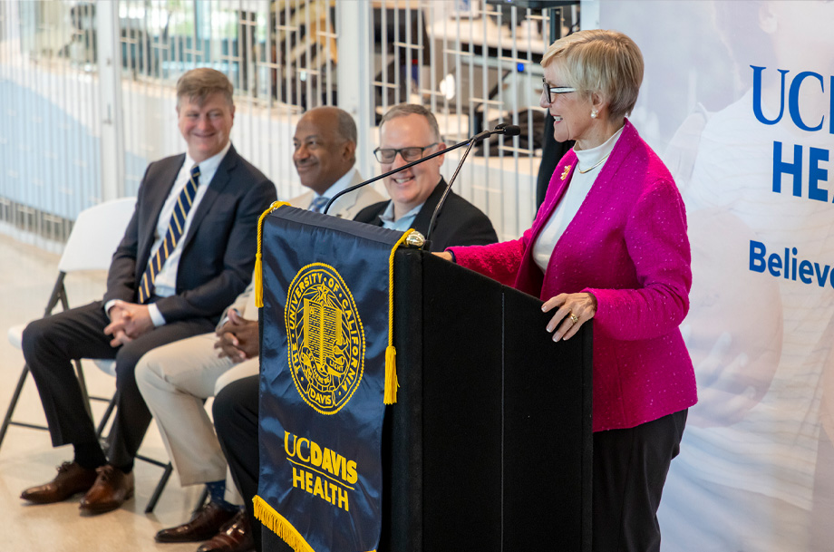 Woman stands at podium draped with UC Davis Health banner speaking into microphone while three other speakers look on