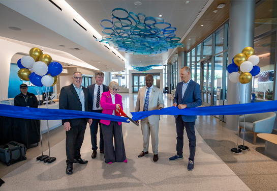 5 hospital leaders stand inside new center each holding part of a blue ribbon while center woman uses large scissors to cut it