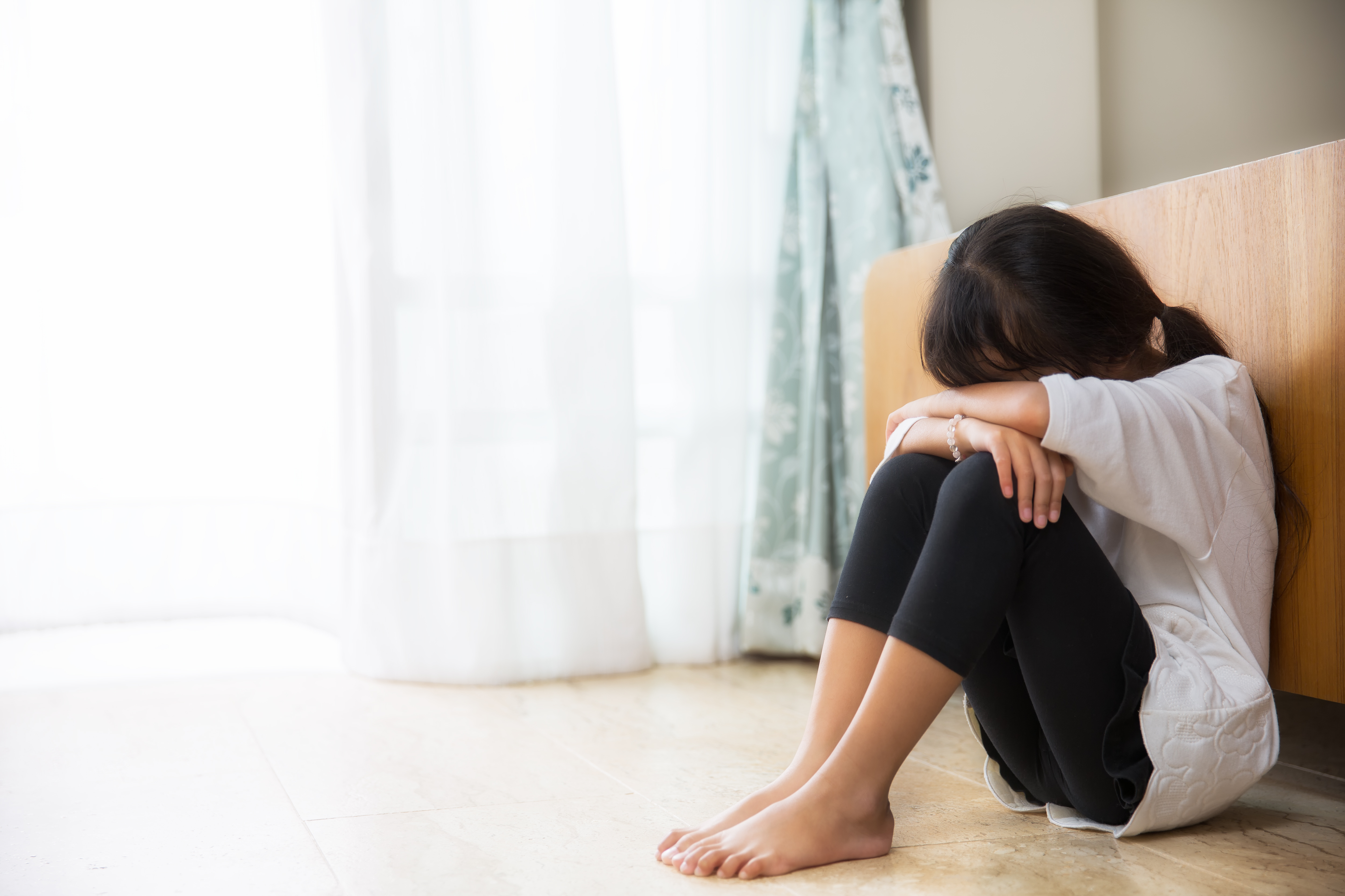 A girl with dark hair and a white T-shirt sits on the floor, leaning her back on a bed frame. Her face is lowered into her arms