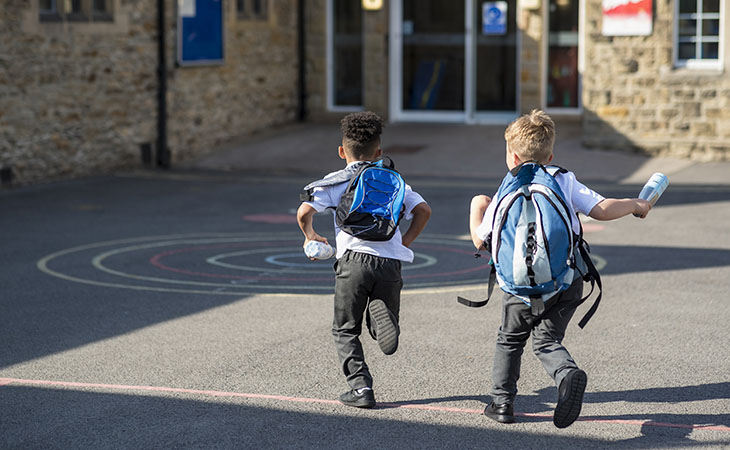 Two elementary school-aged boys run toward a school building, wearing backpacks.