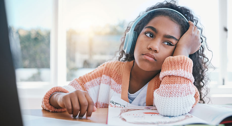 A girl wears headphones while sitting at a desk looking bored