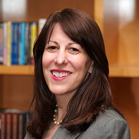 A woman with dark brown hair, wearing a suit, stands near a bookshelf.