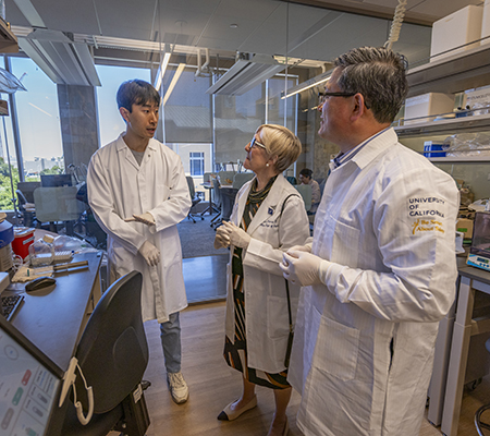 Dr. Diana Farmer standing between Dr. Aijun Wang and a researcher in Wang&rsquo;s Lab. Everyone is wearing a white lab coat.