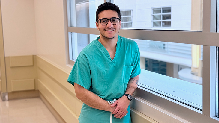 Medical student with dark hair, eyeglasses, green scrubs and tattoo on his left arm leans against a window in a hospital hallway