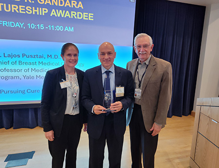 Man holds an award on a stage standing in between a woman with dark ponytail and a man with white hair.