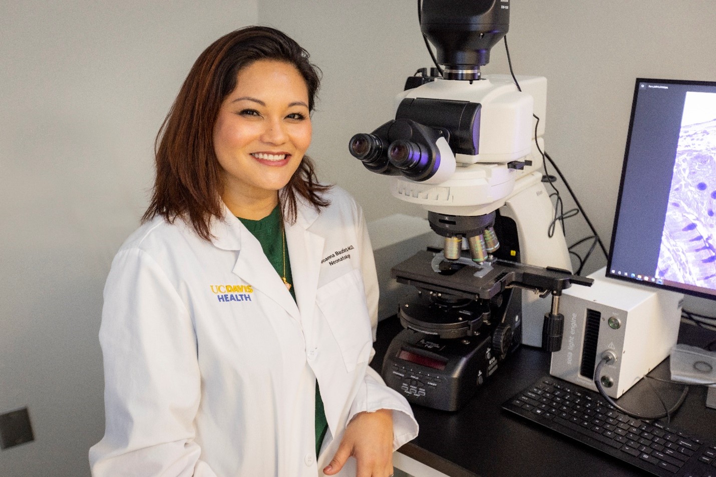 A smiling woman wearing a white lab coat, sitting in front of an electronic microscope, a computer, and a monitor.