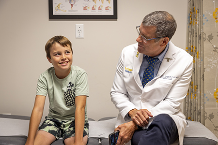 Doctor in white coat talking to a young boy in a green shirt seated on an exam table.
