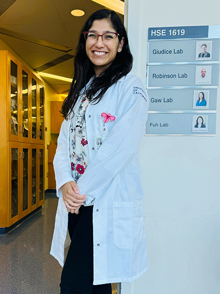 A young woman with dark hair and a white lab coat stands in a hallway next to a small directional sign that states &ldquo;Giudice Lab&rdquo;
