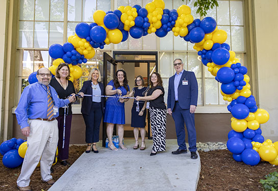 A group of people gather under a blue and gold balloon arch for a ribbon cutting