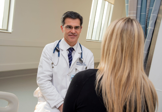 Martin Cadeiras with stethoscope around his neck sitting and speaking to patient with back turned. 