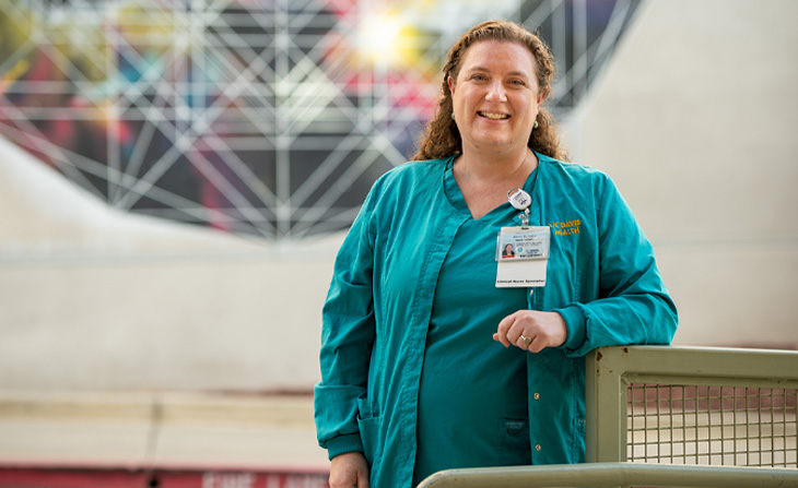 A nurse in teal scrubs smiling in front of a colorful mural.