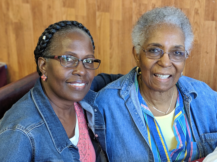 Two women smiling together, sitting close and wearing glasses and denim jackets.