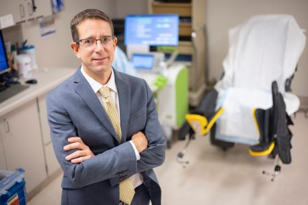 Man with light brown hair in suite with arms crossed standing in exam room.