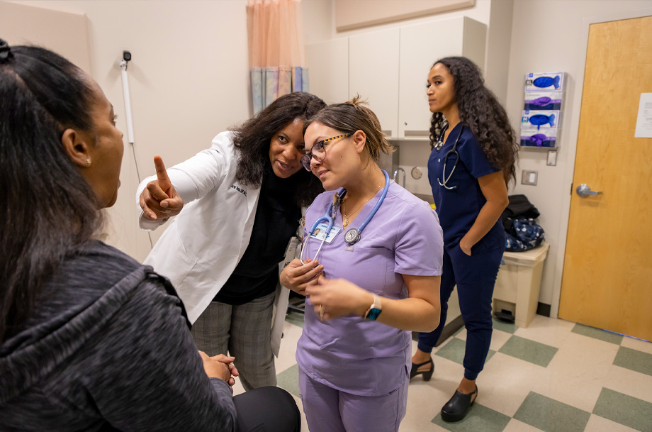 Instructor wearing white coat points at patient in exam room while student with stethoscope around her neck listens