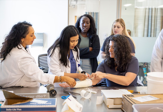 Student wearing white coat practices splinting in skills lab while professor and three other students look on