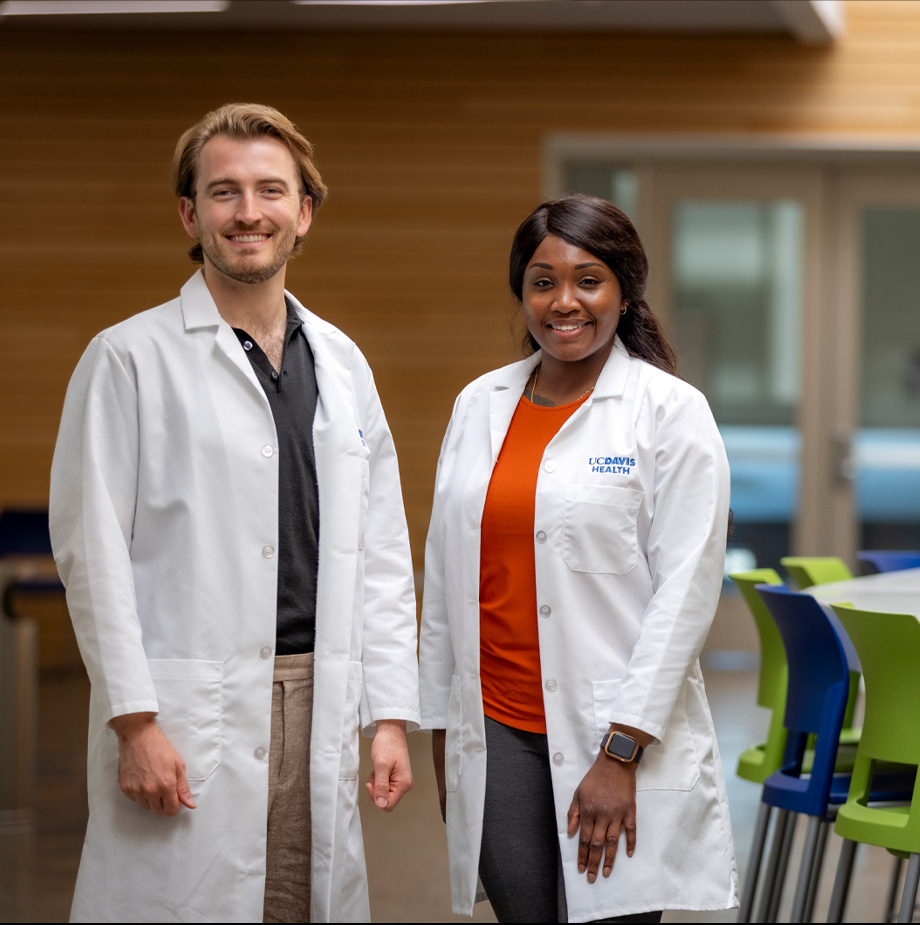 Man on left stands next to woman on right both wearing white coats