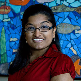 An adult with long dark hair stands before a colorful sea-themed mosaic artwork. 