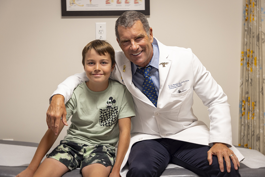 Doctor in white coat with his arm around young boy with green shirt sitting on exam table.