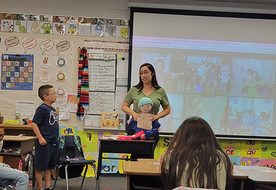 A woman stands in front of the classroom holding a doll. A boy stands to the left of her. 
