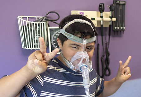 Teenage boy in striped shirt gives a double peace sign as he gets his CPAP adjusted.