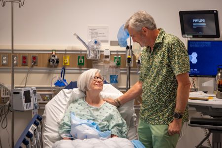 A man stands over a woman's hospital bed, his hand on her shoulder.