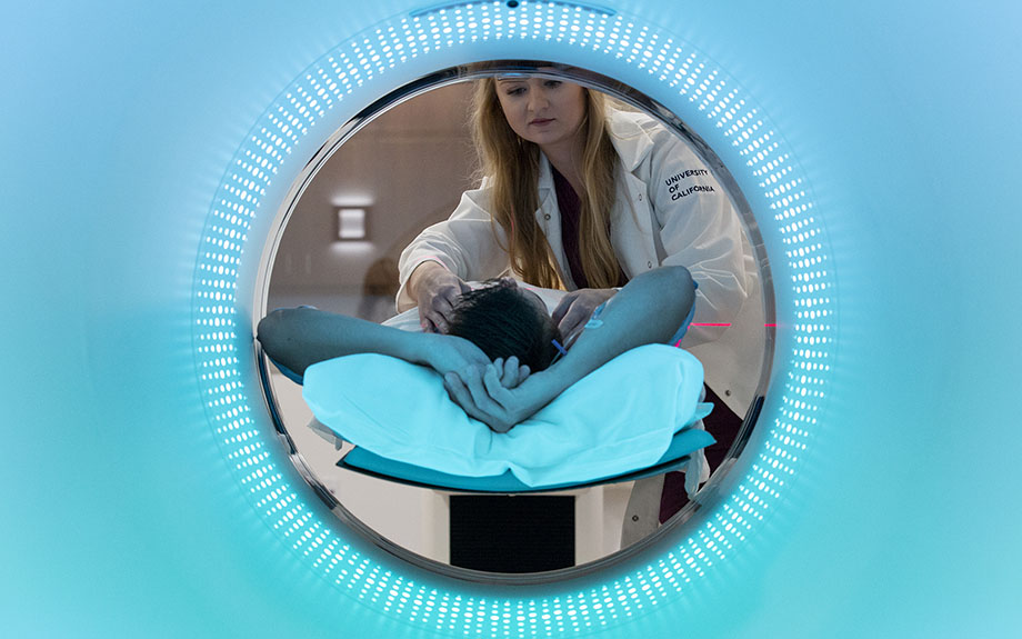 A patient lies on a CT scanner platform while a health care provider helps them. 