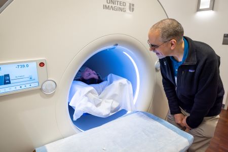 Back of head of man with blue coat watching woman with dark hair enter large imaging machine.