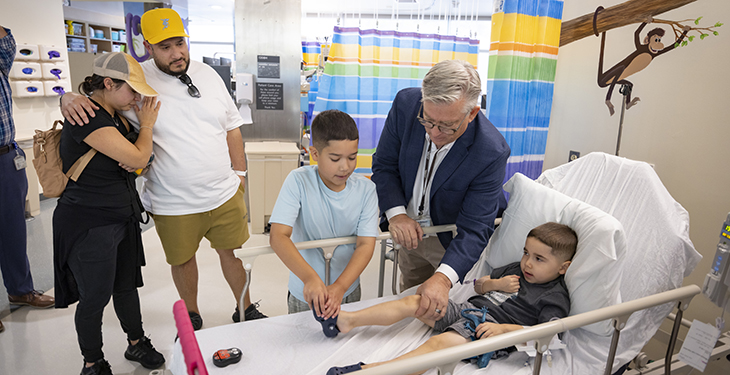 Five year old boy on a hospital bed with Dr. Craig McDonald on his right holding his knee and his brother touching his right toes. Father and mother are in the background.