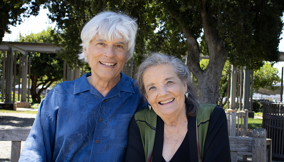 An older man and woman with whitish-gray hair, sit on a wooden bench outdoors. 