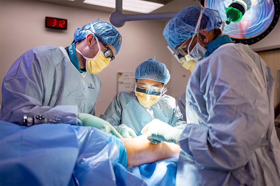 Three people in medical scrubs perform a procedure on a patient&rsquo;s knee in an operating room. 