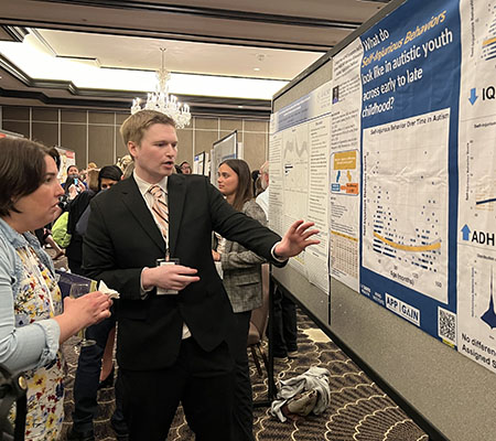 A man in a suit gestures toward a poster presentation at an academic conference.