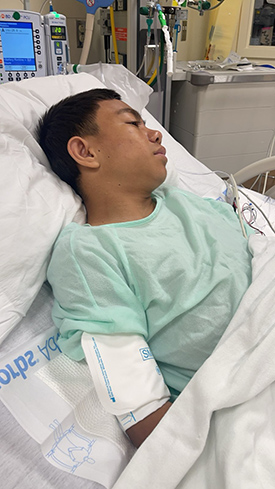 Hispanic teen boy lying in hospital bed with a green gown on.