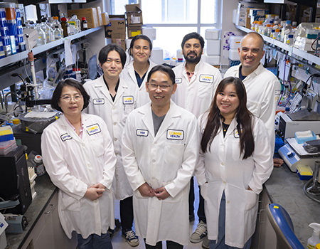 Man in white coat standing in a lab surrounded by six lab members also in white coats.