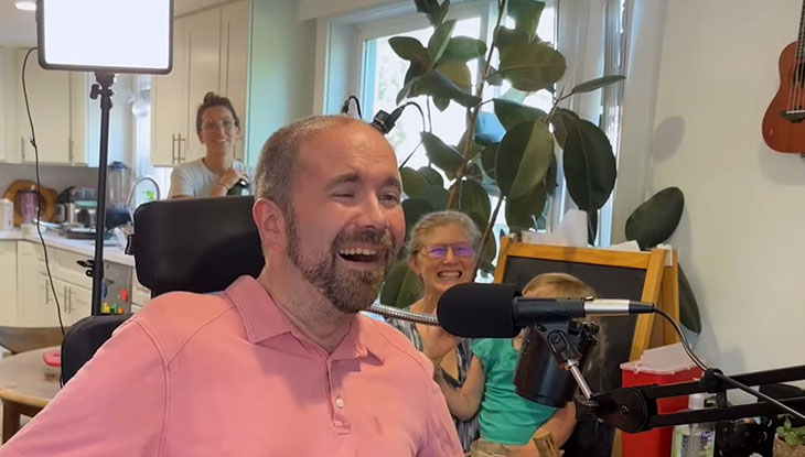 A man in a pink shirt, in a wheelchair, smiles before a computer, surrounded by family.