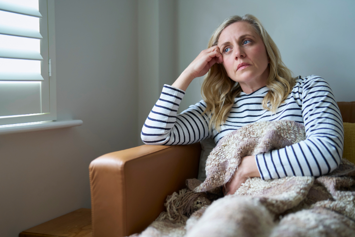 Mature woman in long-sleeve shirt rests her arm on a sofa and holds a lap blanket while looking pensively into the distance