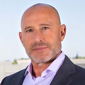 A man wearing a light purple button-down shirt and gray suit jacket poses for a portrait.