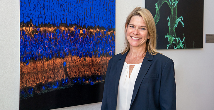 Smiling woman in blazer stands beside colorful microscopic image artwork displayed on a wall.