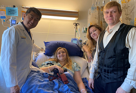 In the center, Tiffany Balts in a hospital bed after her surgery. Neurosurgeon Kee Kim standing to Tiffany’s right and her mother and dad to her left.