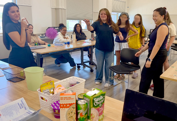 A girl throws a purple squishy ball into a bucket on a table while nursing students and fair participants watch and smile