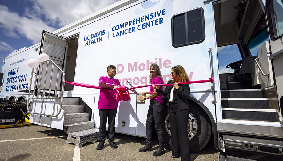 Three adults stand in front of a large mobile mammogram unit to cut a large pink ribbon. 