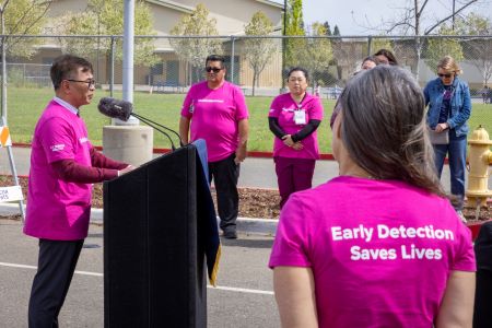 Man with dark hair at podium with pink shirt that reads "MobileMammo+" and others in the audience with pink shirts that read "Early Detection Saves Lives."
