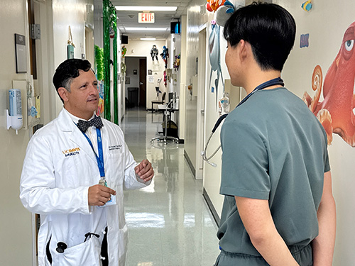 A man with short, dark hair, wearing a purple bow tie and purple shirt, with stethoscope around his neck.