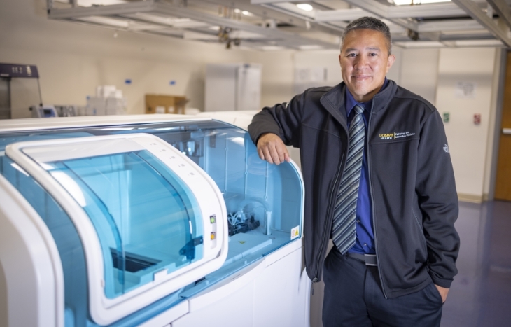 A man in a dark jacket and striped tie stands with his elbow on a beige diagnostic instrument with clear blue windows.