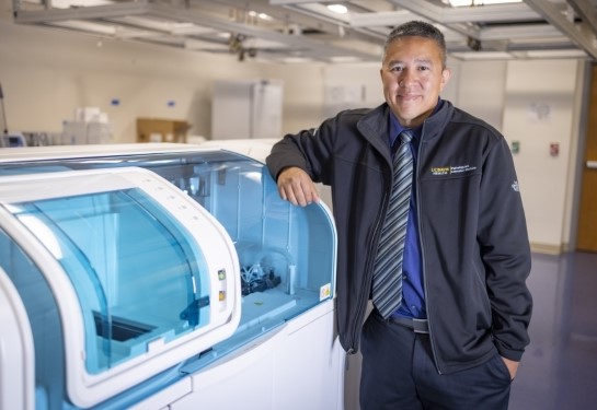 A man in a dark jacket and striped tie stands with his elbow on a beige diagnostic instrument with clear blue windows.